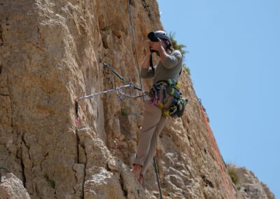 A rock climber takes photos while scaling a rugged cliff under clear blue skies.