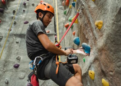 From below side view of attentive young Asian climber in protective helmet using electric screwdriver on hold of artificial rock